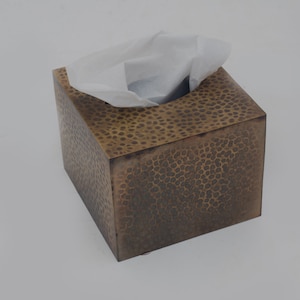Puede incluir: Un porta pañuelos cuadrado de latón martillado con una superficie texturizada. La caja está vacía y tiene un pañuelo blanco asomando por la abertura.