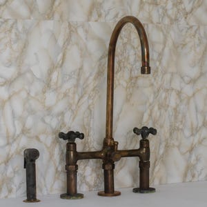 Op de afbeelding: Een bronzen keukenkraan in vintage-stijl met een hoge booguitloop en kruishendelkranen. De kraan heeft een klassiek ontwerp met een aparte zijsproeier. De armatuur staat tegen een marmerpatroonwand.