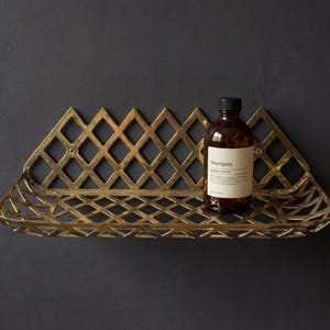 Puede incluir: Estantería de pared de metal dorado con diseño de celosía de diamantes. Una botella de champú marrón con tapa negra y etiqueta blanca descansa sobre la estantería. La estantería está montada en una pared gris oscura.