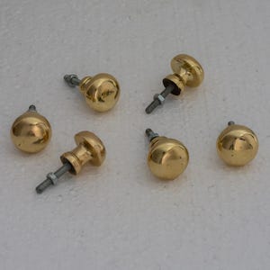 May include: Six brass-colored cabinet knobs with attached screws and nuts. The knobs have a rounded, mushroom-like shape, and are arranged on a white surface. The hardware is likely for furniture restoration or new construction.
