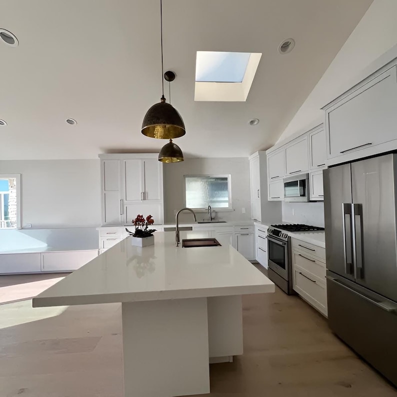 Puede incluir: Una cocina moderna con gabinetes blancos, una isla grande con una encimera blanca y electrodom&eacute;sticos de acero inoxidable. La cocina tiene un tragaluz y dos l&aacute;mparas colgantes doradas sobre la isla.