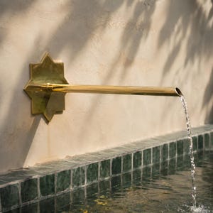 Könnte beinhalten: Ein wandmontierter Brunnenauslauf aus poliertem Messing mit einer sternförmigen Rückplatte. Das lange, zylindrische horizontale Rohr ragt aus der Mitte heraus und leitet einen stetigen Wasserstrahl nach unten in ein gefliestes Becken.
