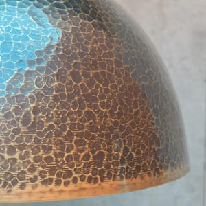 Puede incluir: Una pantalla de l&aacute;mpara de metal martillado con un acabado degradado azul y marr&oacute;n. La pantalla tiene una superficie texturizada y una forma redondeada.