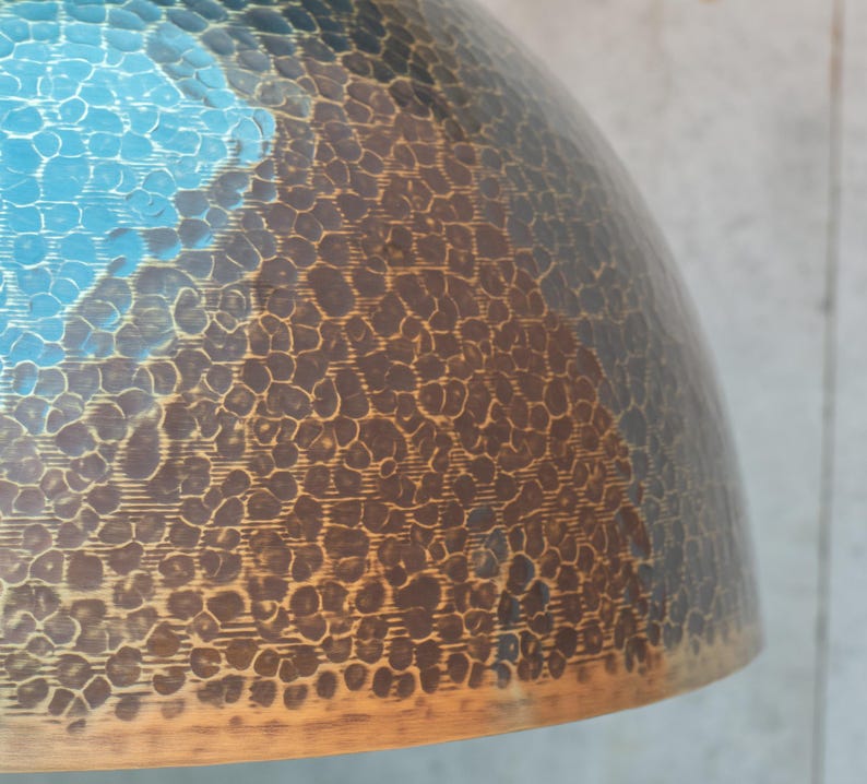 Puede incluir: Una pantalla de l&aacute;mpara de metal martillado con un acabado degradado azul y marr&oacute;n. La pantalla tiene una superficie texturizada y una forma redondeada.