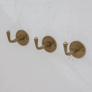 May include: Three brass wall hooks with a curved design are mounted on a white textured wall. Each hook has a circular base and is secured with screws. The hooks are evenly spaced and project outward.