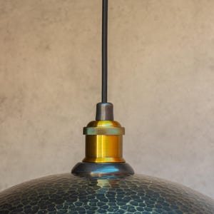 Puede incluir: Una l&aacute;mpara colgante negra y dorada con una pantalla de metal martillado. La luz tiene un cable negro y una toma de corriente dorada.