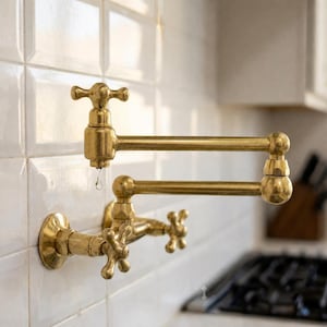Op de afbeelding: Een aan de muur gemonteerde koperen pannenvul-kraan met een dubbel scharnierende arm en twee kruisvormige handgrepen. Het metalen armatuur heeft een gepolijste afwerking en is bevestigd aan een wit betegelde muur, met een waterdruppel zichtbaar.