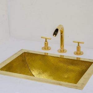May include: A gold hammered brass sink with a rectangular shape and rounded corners. The sink has a single faucet and two handles.