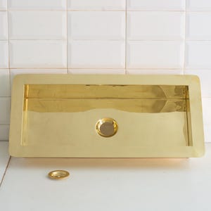 May include: A rectangular, gold-colored sink with a central drain and a matching drain cover. The sink has a polished finish and is set against a white tiled wall. The sink's dimensions are approximately 30 inches long and 15 inches wide.