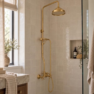 May include: A wall-mounted shower system in a brushed gold finish featuring a round overhead shower head, a vertical pipe, a handheld shower attachment with a flexible hose, and two cross-handle valves mounted on a light beige tiled shower wall.