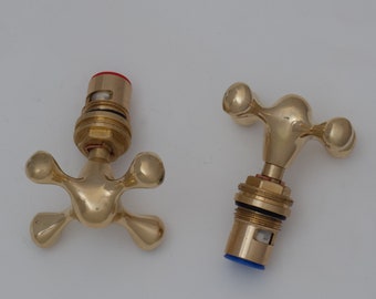 May include: Two polished brass faucet handles with a cross-shaped design. One handle features a red cap on the threaded base, while the other handle features a blue cap on the threaded base. Both handles have four rounded ends and a metallic finish.