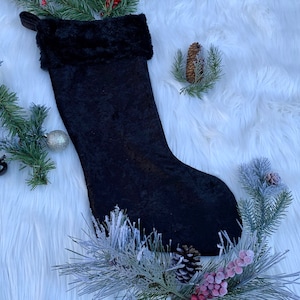 May include: Black velvet Christmas stocking with a faux fur cuff. The stocking is lying on a white fluffy surface with pine sprigs and berries.