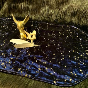 May include: A dark blue velvet table runner with a constellation pattern in gold. A brass sculpture of an animal head, a bone, and two feathers are on the runner. The background is a dark, furry material.