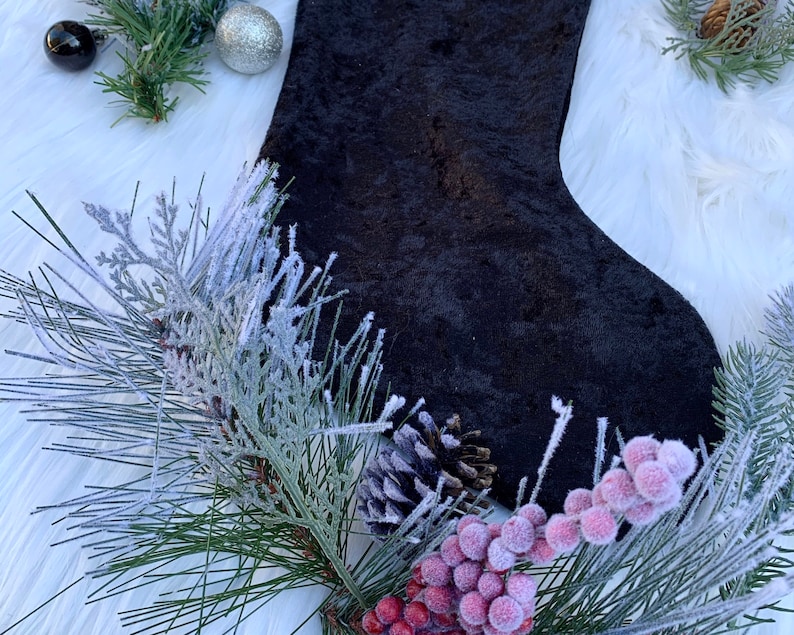 May include: A black velvet stocking with a white fluffy background. The stocking is decorated with a pine sprig and red berries.