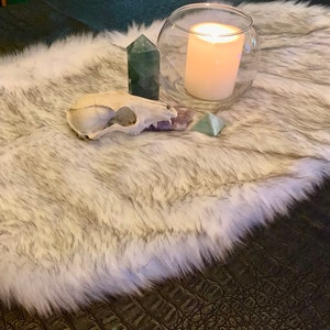 May include: A white faux fur rug with a white animal skull, a green crystal, a purple crystal, and a clear glass jar with a white candle inside.