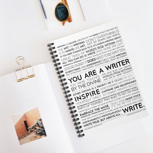 May include: A black and white notebook with a spiral binding. The cover features a motivational quote about writing, including the words "You are a writer" and "Inspire yourself daily".