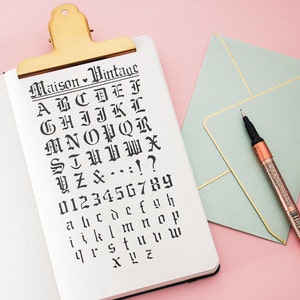 Könnte beinhalten: Ein weißes Notizbuch mit einem goldenen Clip, geöffnet auf einer Seite mit einem handgeschriebenen Alphabet und Zahlen in einer Vintage-Schriftart. Das Notizbuch liegt auf einer rosa Oberfläche neben einem hellgrünen Umschlag mit goldener Umrandung und einem goldenen Stift.