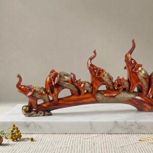 May include: A wooden sculpture of a family of elephants. The elephants are a reddish-brown color with gold accents. The sculpture is displayed on a white marble surface.