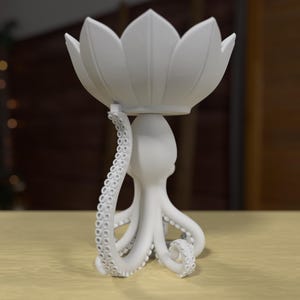 以下が含まれることがあります： 白い装飾彫刻。蓮の花のボウルがタコによって支えられています。タコには吸盤のある複数の触手があります。蓮の花には複数の花びらがあります。彫刻は薄い黄色の表面にあります。