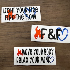May include: Three white stickers with black text and orange and blue flames and hearts. The first sticker says "LIGHT YOUR FIRE FIND THE FLOW". The second sticker says "F&F" with a flame on the left and a heart on the right. The third sticker says "MOVE YOUR BODY RELAX YOUR MIND".