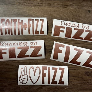 May include: Five white vinyl stickers with rose gold glitter lettering. The stickers feature the text "Faith + Fizz", "Fueled by Fizz", "Running on Fizz", "Fizz" and "Peace Love Fizz".