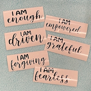 May include: Six white stickers with black lettering. Each sticker has the phrase "I AM" followed by a different word: "enough", "empowered", "driven", "grateful", "forgiving", and "fearless".