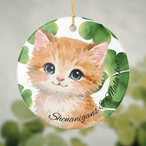 May include: Round ceramic ornament featuring a watercolor illustration of a ginger kitten with blue eyes. The ornament is decorated with green shamrocks and the words "Shenanigans & Malarkey". A gold string is attached for hanging.