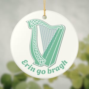May include: A round, white ceramic ornament with a gold string for hanging. The ornament features a green harp design with the words "Erin go bragh" in green script. The harp has intricate Celtic knot detailing.