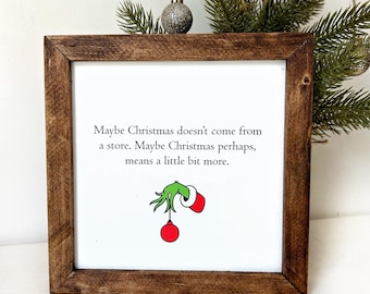 Puede incluir: Una impresión enmarcada con fondo blanco y marco marrón. La impresión presenta una mano verde del Grinch sosteniendo un adorno rojo y el texto "Maybe Christmas doesn't come from a store. Maybe Christmas perhaps, means a little bit more."