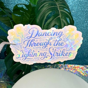 May include: A holographic sticker with the text "Dancing Through the Lightning Strikes" in blue script. The sticker features lightning bolt and star designs. The sticker is held by a pair of silver tweezers, with green leaves in the background.