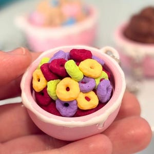 May include: A miniature pink bowl filled with colorful cereal, held in a hand. The cereal pieces are various colors, including yellow, purple, red, and green. Two other miniature bowls with cereal are in the background, on a white cake stand.