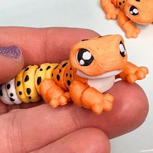 Puede incluir: Tres figuras de gecko naranja y amarillo con manchas negras y ojos grandes y expresivos. Las figuras de gecko están sobre un plato blanco. Una persona sostiene un gecko. Las figuras de gecko tienen un diseño lindo y caprichoso.