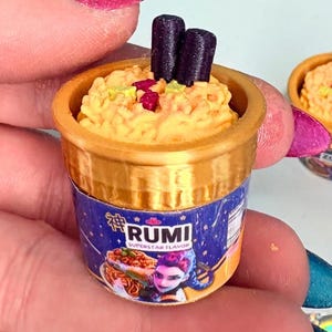 May include: Miniature ramen cup with a gold rim and a blue label that reads "RUMI". The cup is filled with yellow noodles, topped with dark brown sticks and red accents. A second, smaller cup is visible in the background.