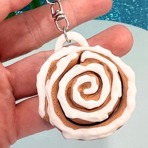 Könnte beinhalten: Ein Zimtschnecken-Schlüsselanhänger mit einem silberfarbenen Metallverschluss. Die Miniaturrolle ist braun mit weißem Zuckerguss und einer Schlaufe oben, die mit dem Verschluss verbunden ist. Der Schlüsselanhänger wird in einer Hand gehalten.