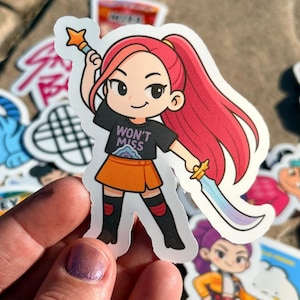 May include: A sticker featuring a cartoon character with red hair, an orange skirt, and a black shirt that says "WON'T MISS". The character is holding a star-shaped wand and a sword. Other stickers are in the background.