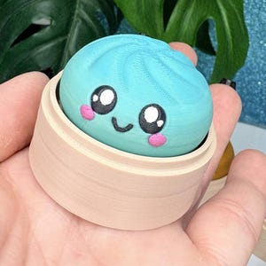 Può includere: Una statuetta a forma di gnocco, arrotondata e testurizzata, di colore azzurro con un viso sorridente, grandi occhi bianchi e neri e guance rosa, posta all'interno di un piccolo contenitore cilindrico color beige chiaro.