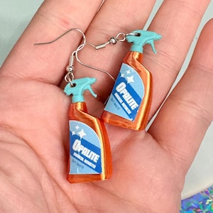 May include: A pair of novelty earrings shaped like miniature orange spray bottles with blue spray nozzles. Each bottle has a blue and white label with the word "Opalite" and a star design. The earrings are held by silver fish hook ear wires.