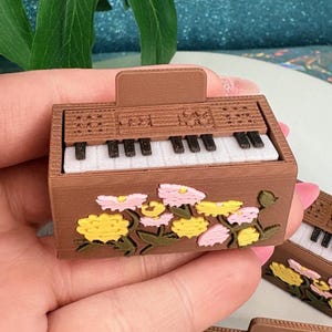 May include: Brown miniature piano with white keys and floral accents in yellow and pink. The small piano has a rectangular shape and is decorated with a floral design on the front. The keys are black and white.