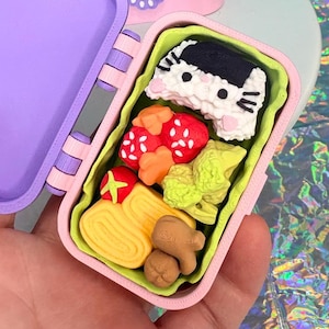 May include: A miniature, open purple and pink bento box filled with colorful, detailed food items. The food includes a cat-shaped sushi, red strawberries, and yellow omelet. Additional closed bento boxes are in the background.
