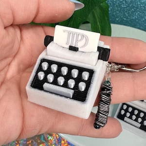 Könnte beinhalten: Ein weißer Miniatur-Schreibmaschinen-Schlüsselanhänger mit schwarzen Tasten und Akzenten. Die Schreibmaschine hat ein kleines Stück Papier mit den Buchstaben "TP". Der Schlüsselanhänger hat einen silberfarbenen Ring und einen dekorativen schwarz-silbernen Anhänger.