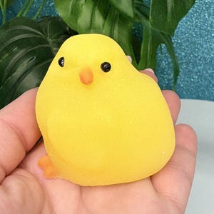 Può includere: Una palla antistress a forma di pulcino giallo brillante, con occhi neri e becco e zampe arancioni. Il pulcino è tenuto in una mano, con uno sfondo di foglie verdi e una superficie blu scintillante.