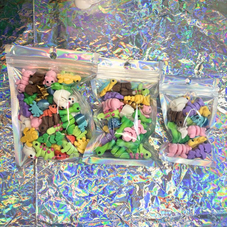 May include: Three clear plastic bags filled with colorful polymer clay animal figurines. The figurines include various colors such as pink, green, yellow, brown, and purple. The bags are sealed and arranged on a shiny, iridescent surface.