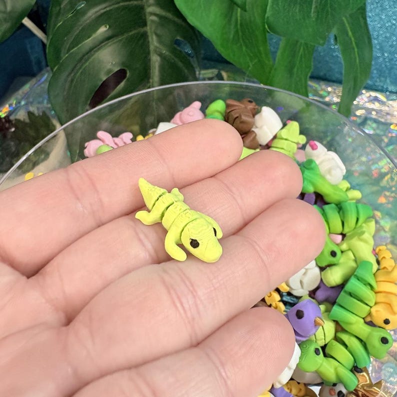 May include: A small, yellow alligator figurine with black eyes is held in a hand. The figurine is surrounded by other colorful polymer clay animal and plant shapes, including green alligators, pink flowers, and brown bears. The items are in a clear glass bowl.