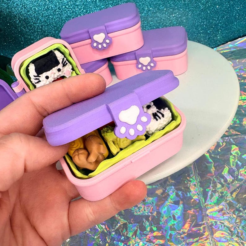 May include: Pink and purple miniature lunch boxes with cat paw closures. One box is open, revealing a miniature cat-shaped food item. The other boxes are closed. The lunch boxes are on a white surface.