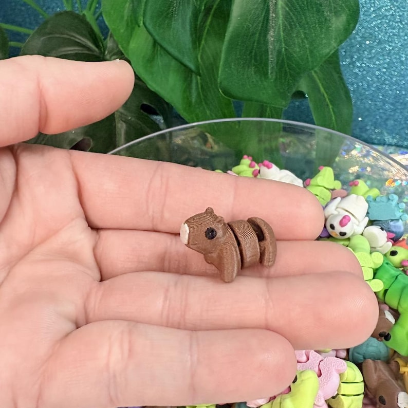 May include: A small, brown capybara figurine with a white muzzle and black eyes is held in a hand. The figurine is segmented, giving it a unique appearance. Other colorful miniature animal figurines are visible in the background.
