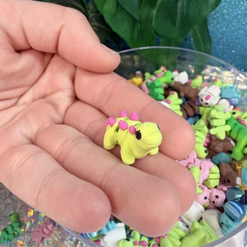 May include: A small, lime green figurine of a cartoon-style dinosaur with pink spikes along its back. The figurine is held in a hand, with other similar figurines visible in the background, suggesting a collection or craft supply.