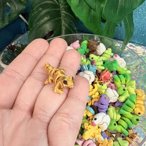 May include: A small, gold-colored lizard figurine with black eyes is held in a hand. The figurine is surrounded by a variety of colorful, miniature clay sculptures of animals and plants in a clear bowl. The background includes green leaves.