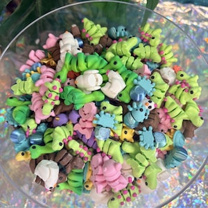 May include: A clear glass bowl filled with colorful polymer clay figurines. The figurines are in various shapes, including caterpillars, flowers, and abstract forms, in shades of green, pink, blue, brown, and white. The bowl sits on a reflective surface.