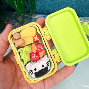 May include: A miniature yellow bento box, open to reveal a variety of food items. The box contains a rolled omelet, a small tomato, broccoli, sushi with a cat face, and other food items. The box is held in a hand.