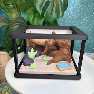 Puede incluir: Un diorama impreso en 3D dentro de un marco rectangular negro que presenta una criatura esculpida marrón con cuernos, rocas geométricas gris oscuro, plantas suculentas verde claro, un pequeño cuenco de agua azul y una base de arena texturizada.